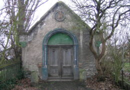 Eine alte Kapelle steht zwischen zwei Bäumen in herbstlicher Atmosphäre.