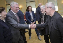 Zwei Männer und zwei Frauen stehen im Vordergrund, die Männer reichen sich die Hand, weitere Personen im Hintergrund