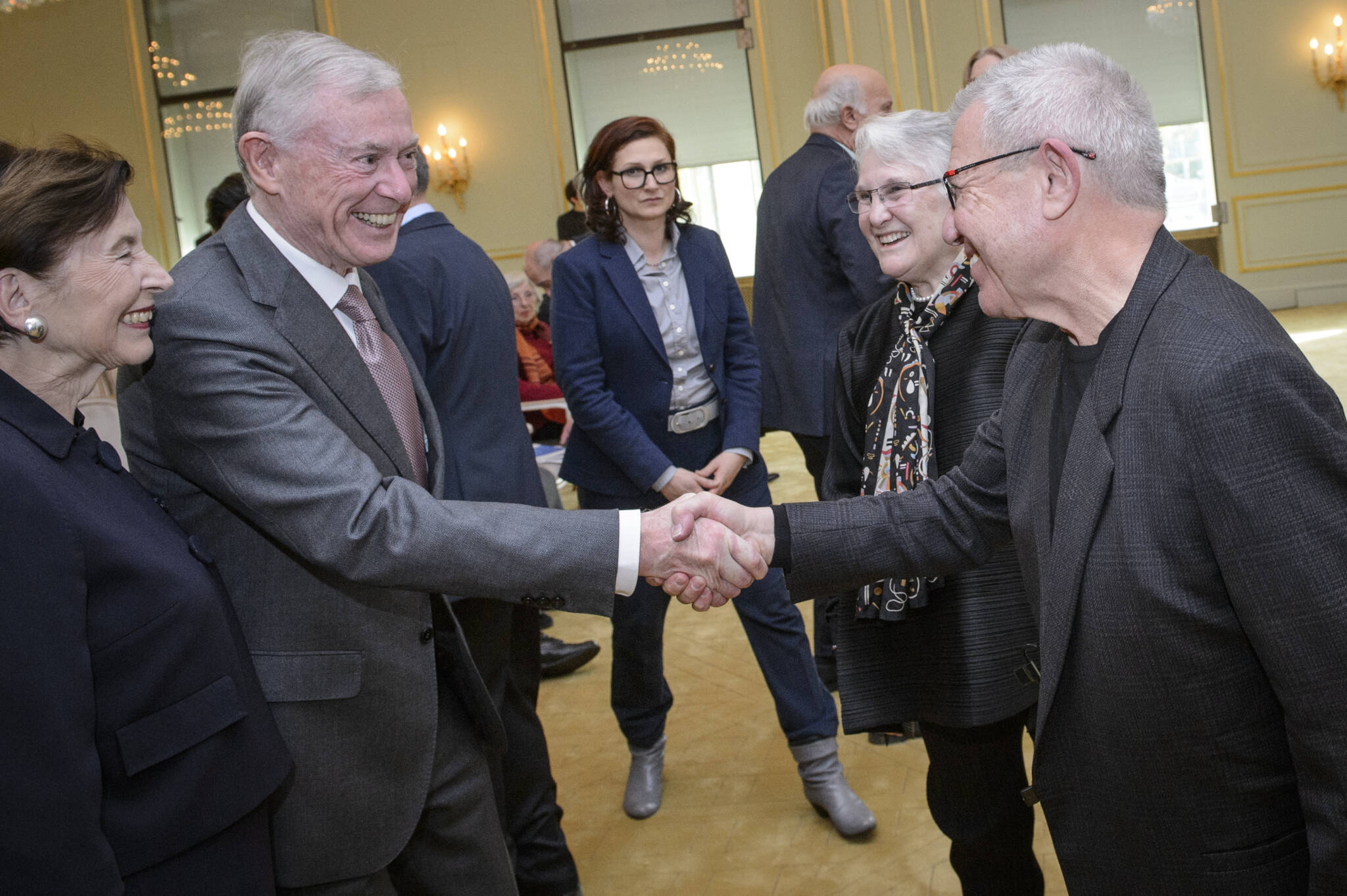 Zwei Männer und zwei Frauen stehen im Vordergrund, die Männer reichen sich die Hand, weitere Personen im Hintergrund